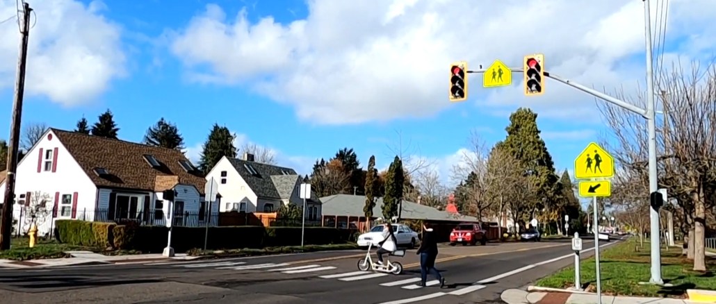 A child on an electric scooter and a pedestrian use a marked crosswalk with a school crossing traffic sign to cross in front of oncoming traffic.