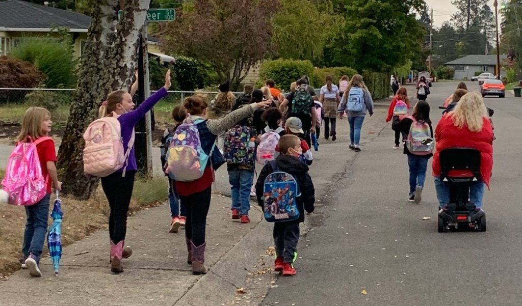 A large group of elementary school students walking to school on the sidewalk and in the street in a residential area on a fall day, with a parent on a scooter accompanying them.